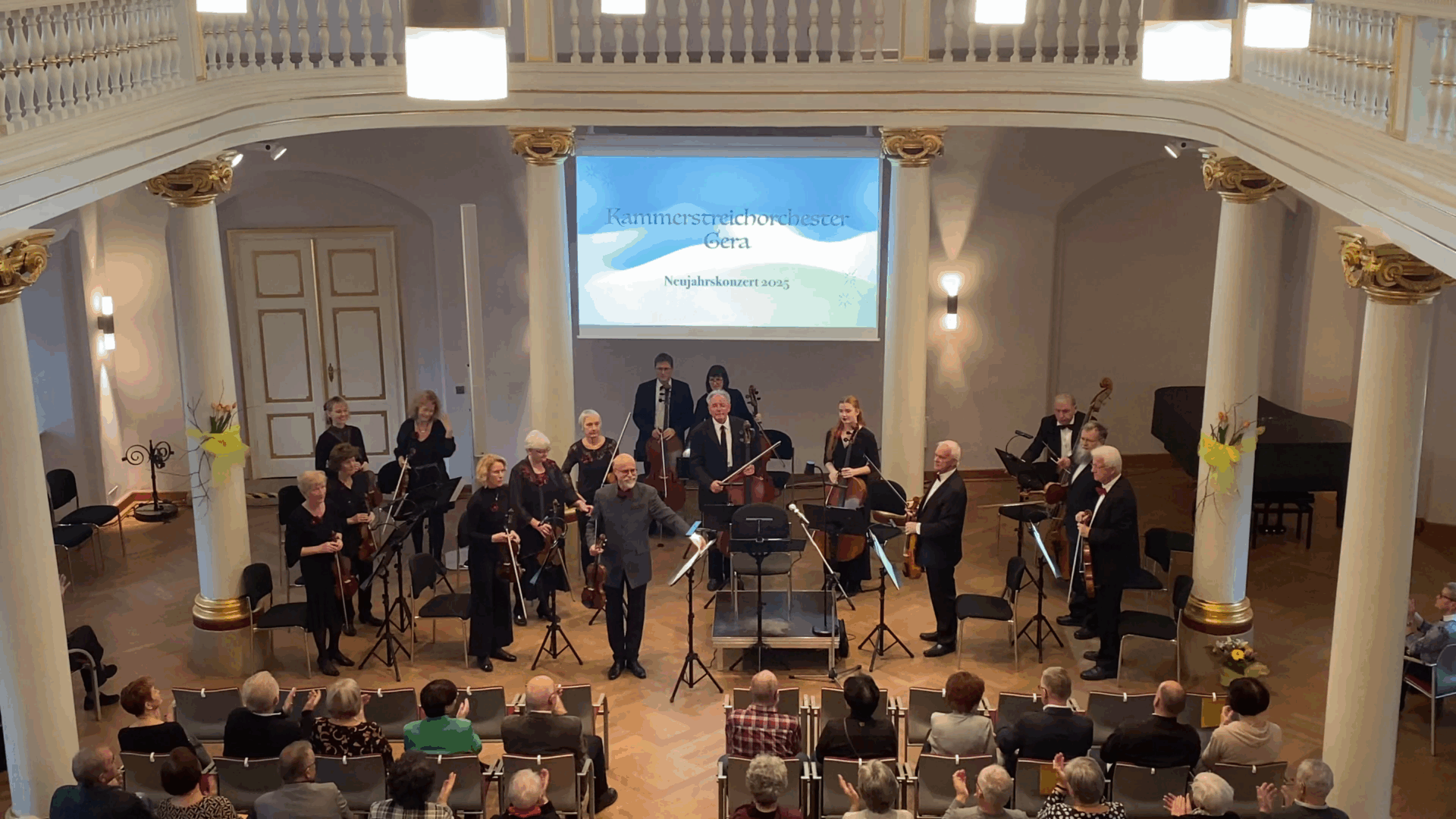 In der Mitte des Bildes steht der Dirigent Peter Wiegand vor dem Orchester, die Arme ausgebreitet, während das Publikum applaudiert. Die Musikerinnen und Musiker, größtenteils in schwarzer Kleidung, stehen vor ihrem Publikum und verbeugen sich nach dem Neujahrskonzert 2025.