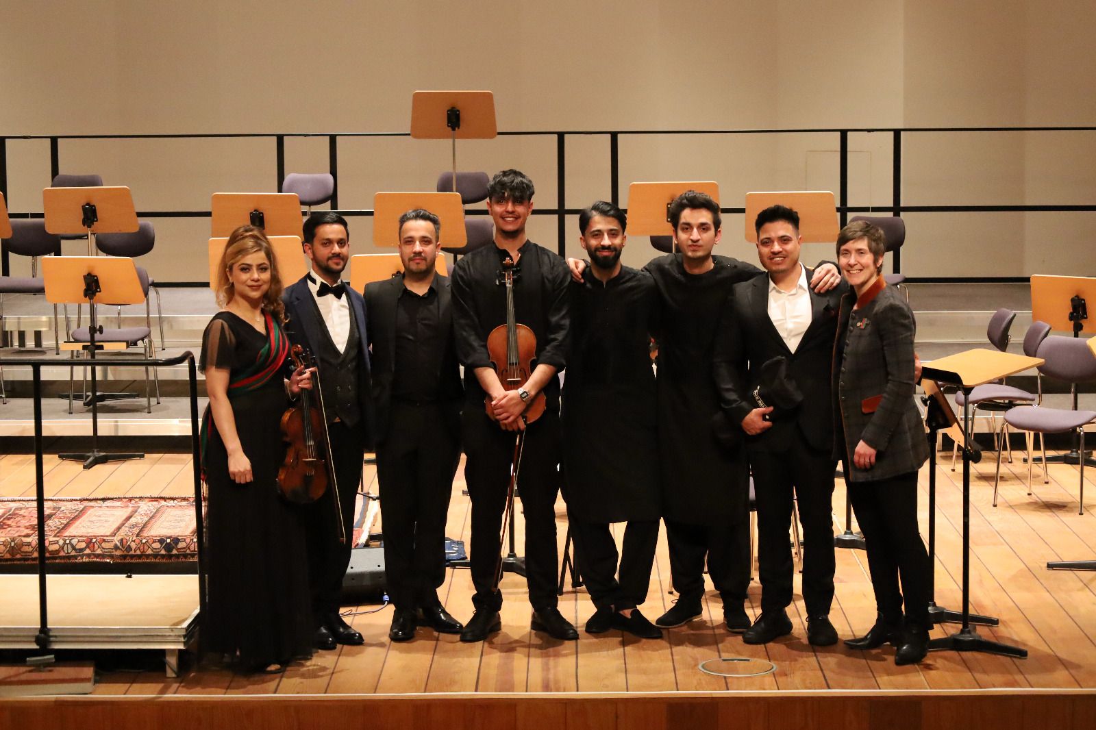 Alle acht afghanischen Mitspieler*innen stehen, teils mit ihren Instrumenen, auf ansonsten leerer Bühne und strahlen.